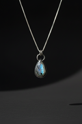 A silver teardrop pendant necklace featuring a shimmering iridescent blue-green stone, suspended from a delicate silver chain against a dark background.