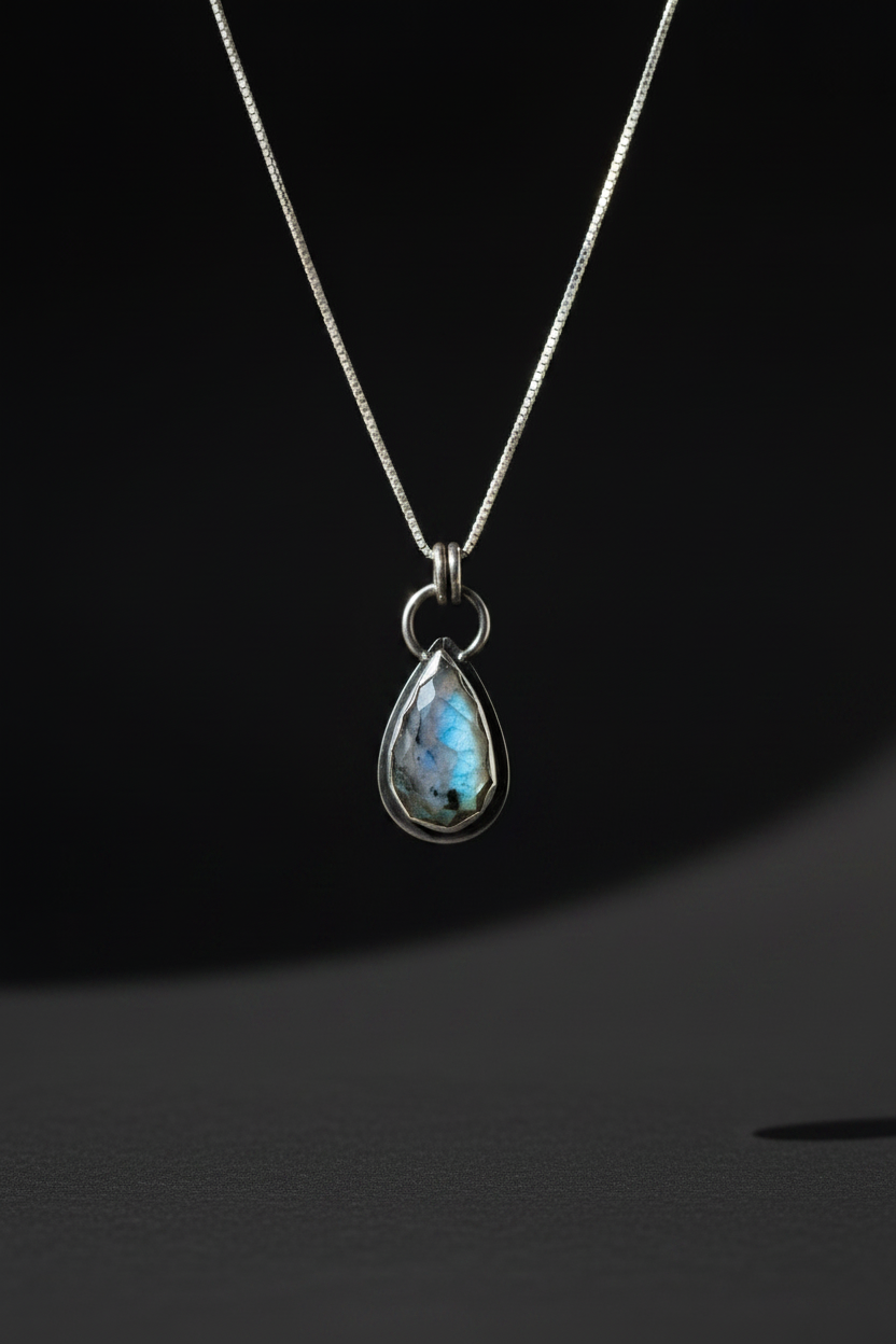 A silver teardrop pendant necklace featuring a shimmering iridescent blue-green stone, suspended from a delicate silver chain against a dark background.