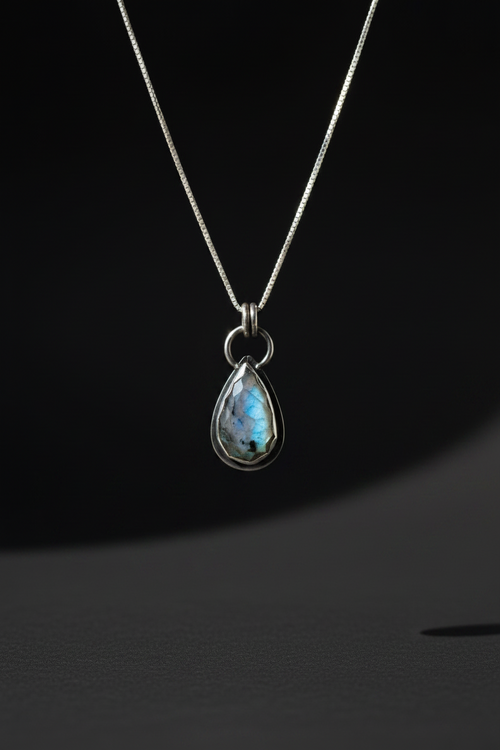 A silver teardrop pendant necklace featuring a shimmering iridescent blue-green stone, suspended from a delicate silver chain against a dark background.