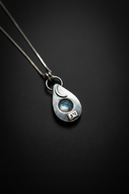 A sterling silver teardrop pendant necklace featuring a crescent moon, a small blue moonstone, and a 'DN' initial charm, shown on a dark background.