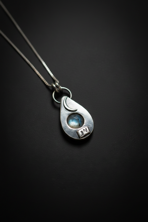 A sterling silver teardrop pendant necklace featuring a crescent moon, a small blue moonstone, and a 'DN' initial charm, shown on a dark background.