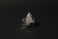 A silver ring featuring a large, teardrop-shaped clear gemstone with black inclusions, set against a dark, reflective background.