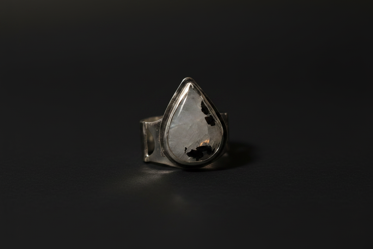 A silver ring featuring a large, teardrop-shaped clear gemstone with black inclusions, set against a dark, reflective background.