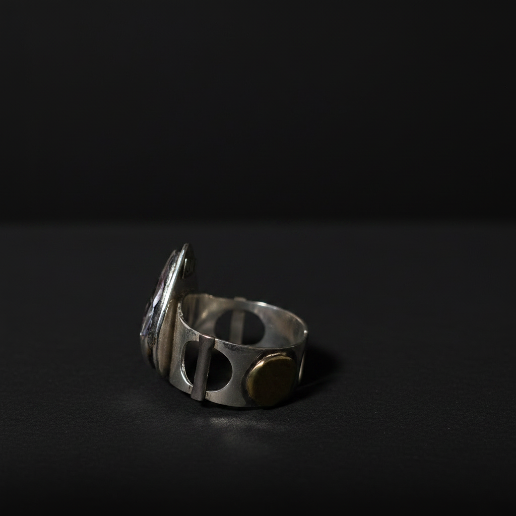 A silver-toned metal ring with a unique slotted band design and a small round gold-toned accent, photographed on a dark black background.