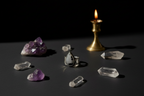 A silver ring with a teardrop-shaped black and white stone is displayed on a dark surface, surrounded by clear quartz crystals, a purple amethyst cluster, and a lit brass candle holder against a black background.