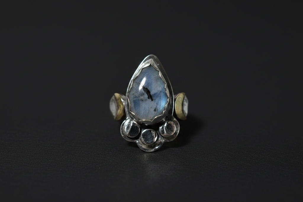 A sterling silver ring featuring a large teardrop-shaped blue labradorite stone in the center, flanked by two small round gold and silver beads, and finished with a cluster of small round clear stones at the base, set against a dark background.