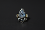 A sterling silver ring featuring a large teardrop-shaped blue labradorite stone in the center, flanked by two small round gold and silver beads, and finished with a cluster of small round clear stones at the base, set against a dark background.