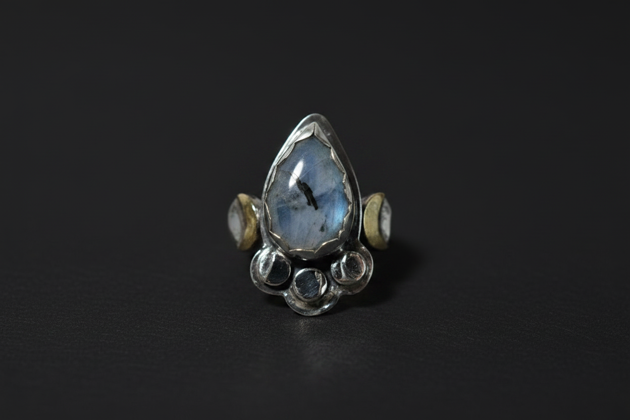 A sterling silver ring featuring a large teardrop-shaped blue labradorite stone in the center, flanked by two small round gold and silver beads, and finished with a cluster of small round clear stones at the base, set against a dark background.