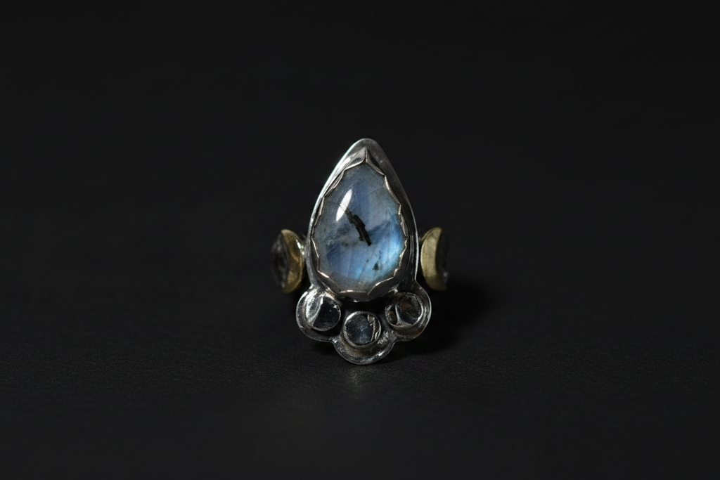 A silver ring featuring a teardrop-shaped blue labradorite stone in the center, flanked by small gold crescent moon accents and circular silver details at the base, set against a dark background.