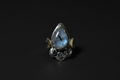 A silver ring featuring a teardrop-shaped blue labradorite stone in the center, flanked by small gold crescent moon accents and circular silver details at the base, set against a dark background.