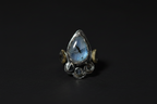 A silver ring featuring a teardrop-shaped blue labradorite stone in the center, flanked by small gold crescent moon accents and circular silver details at the base, set against a dark background.