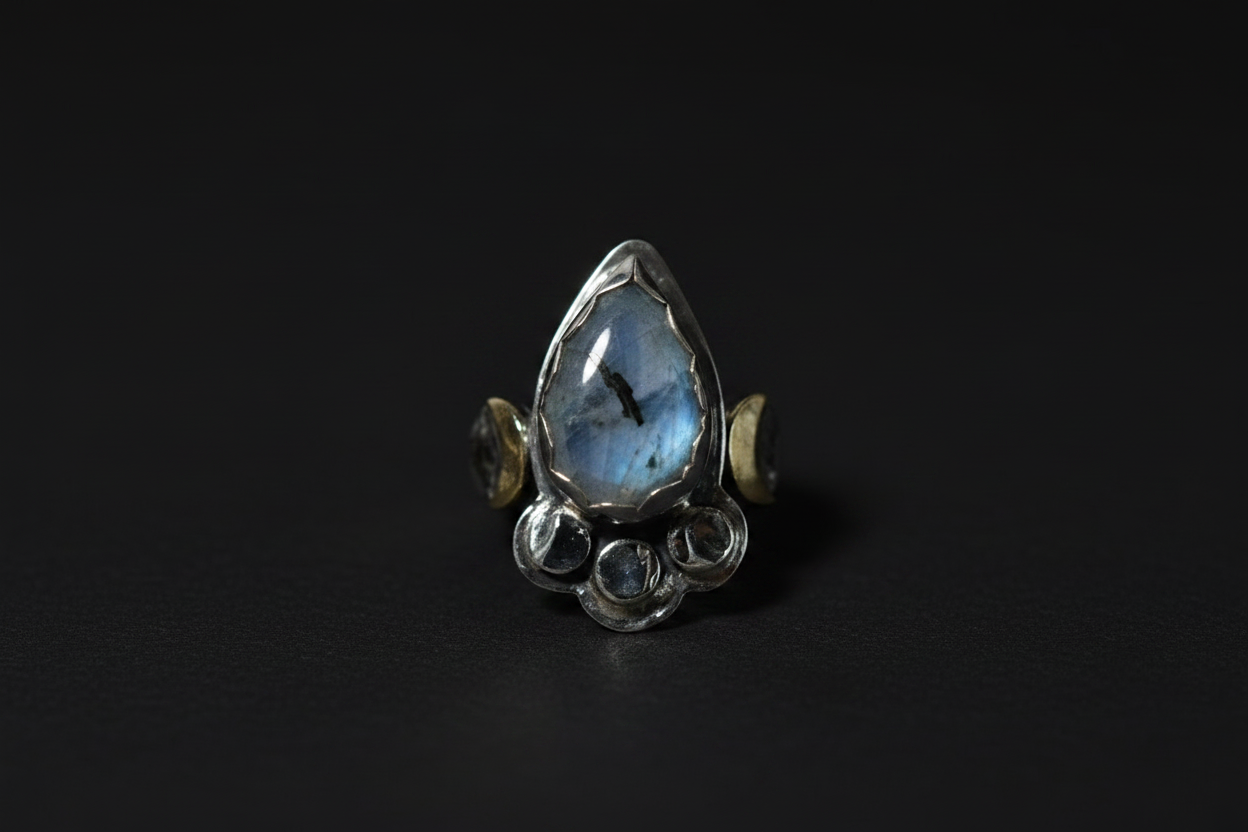 A silver ring featuring a teardrop-shaped blue labradorite stone in the center, flanked by small gold crescent moon accents and circular silver details at the base, set against a dark background.