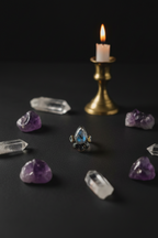 A silver ring with a teardrop-shaped blue gemstone is displayed on a dark surface, surrounded by raw purple amethyst and clear quartz crystals, with a lit brass candle holder in the background.