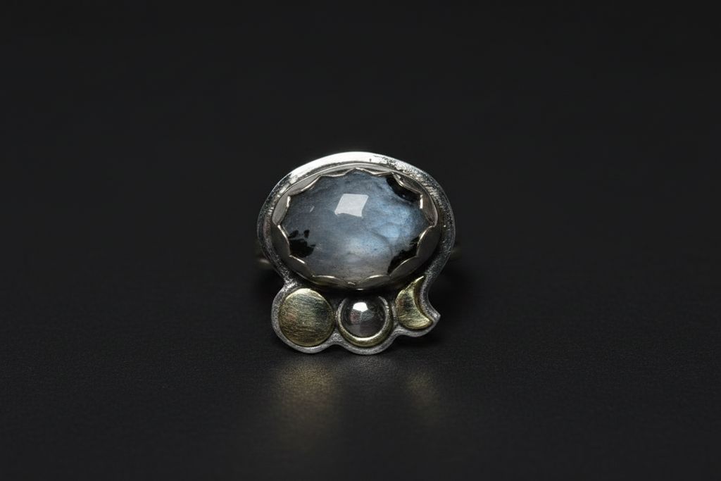 A sterling silver ring featuring a large, round, iridescent gray-blue stone with black inclusions, accented by three smaller silver beads along the band, set against a dark black background.