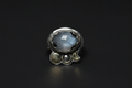 A sterling silver ring featuring a large, round, iridescent gray-blue stone with black inclusions, accented by three smaller silver beads along the band, set against a dark black background.