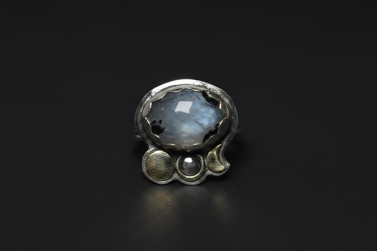 A sterling silver ring featuring a large, round, iridescent gray-blue stone with black inclusions, accented by three smaller silver beads along the band, set against a dark black background.