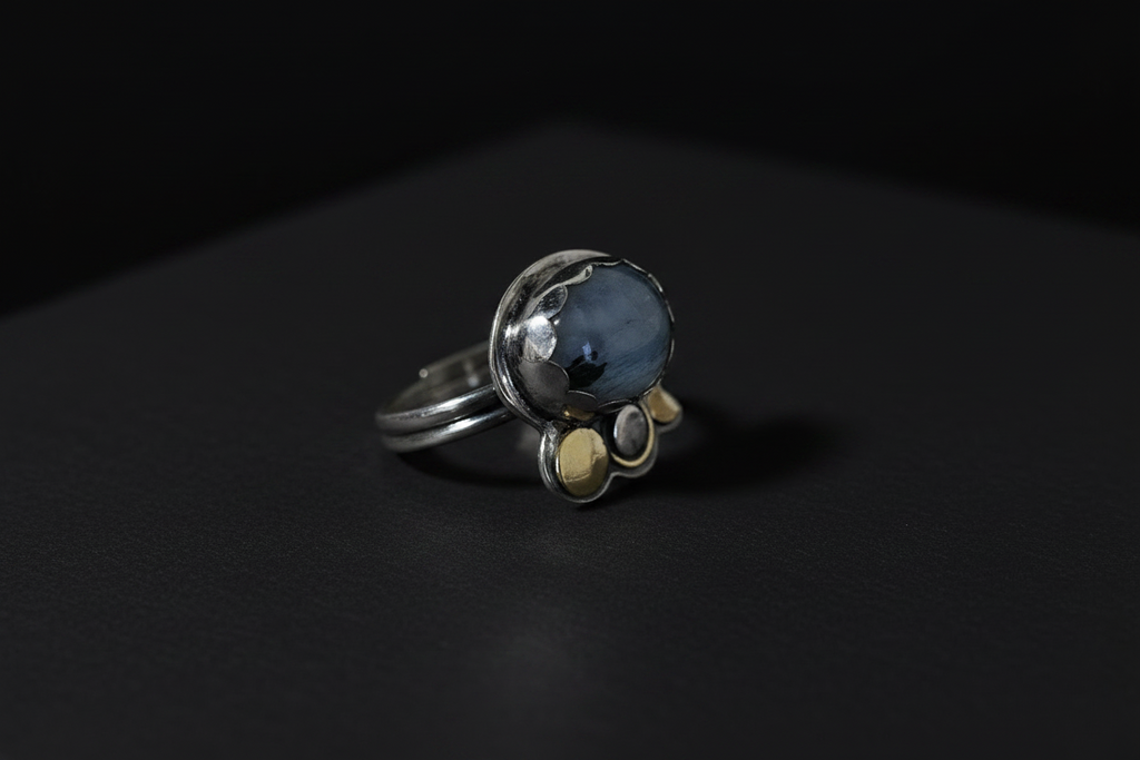 A silver ring with a round blue-gray stone and three small gold accent stones, photographed on a dark black background.