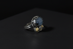 A silver ring with a round blue-gray stone and three small gold accent stones, photographed on a dark black background.