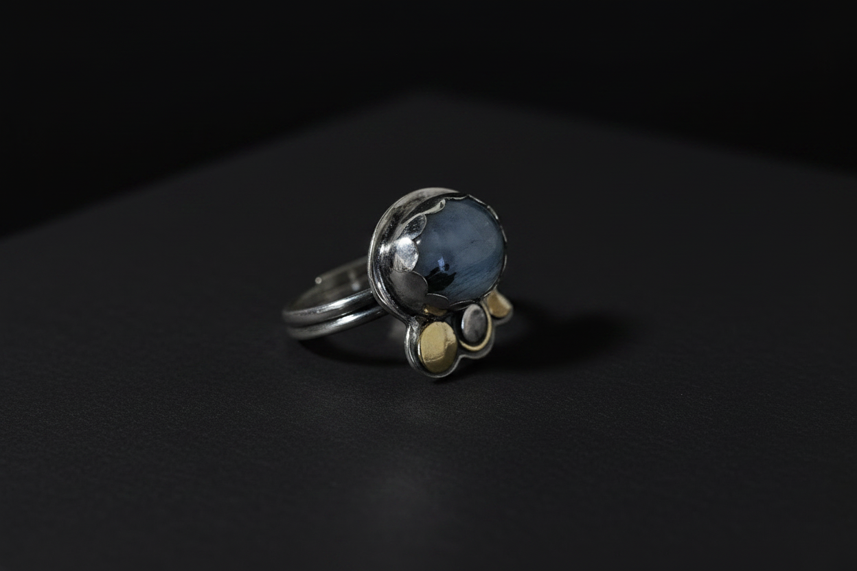 A silver ring with a round blue-gray stone and three small gold accent stones, photographed on a dark black background.