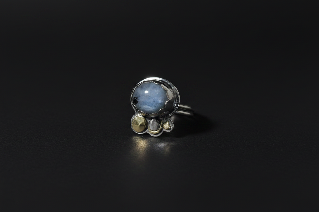 A silver ring featuring a large oval light blue stone with smaller round clear and yellowish-green stones clustered beneath it, set against a dark black background.