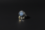 A silver ring featuring a large oval light blue stone with smaller round clear and yellowish-green stones clustered beneath it, set against a dark black background.