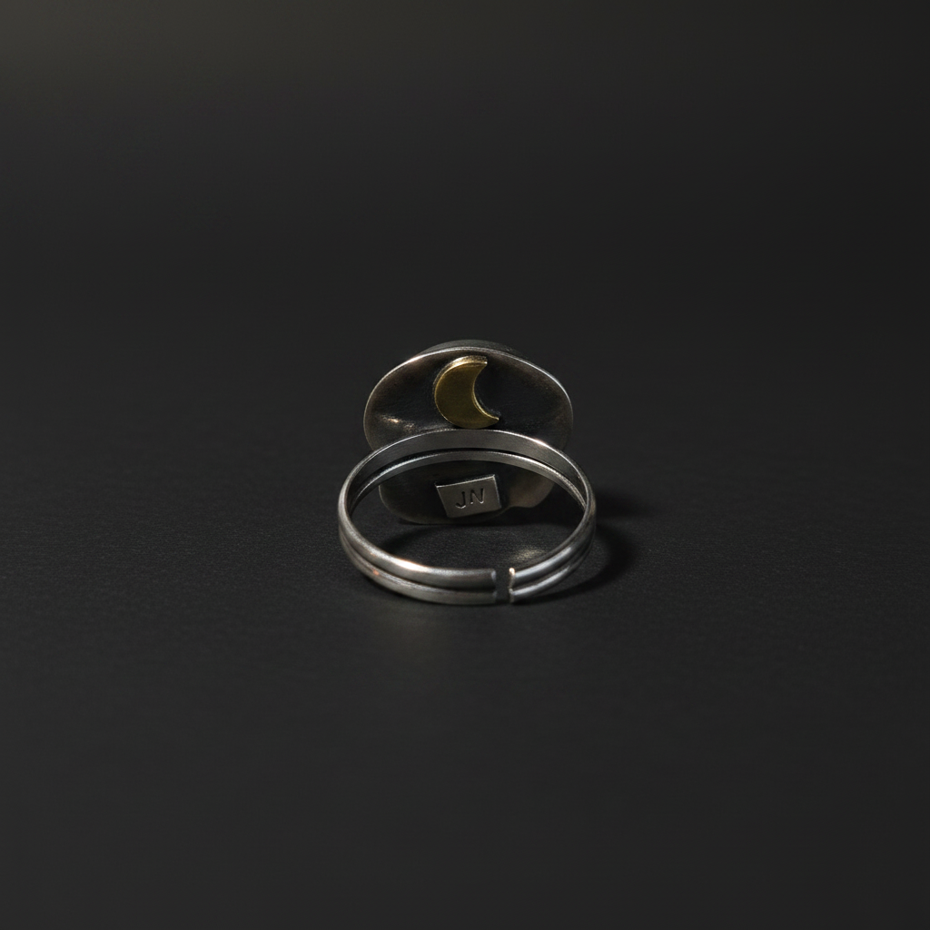 Two silver-toned open stretch rings stacked together on a dark black background. The top ring features a small gold crescent moon inlay, and the bottom ring has a small square brand plaque inside.