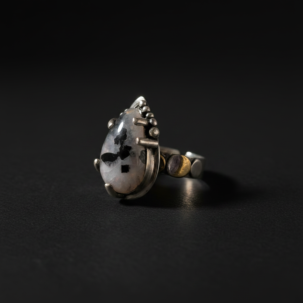 A silver ring featuring a teardrop-shaped white stone with black speckles, set against a dark black background.