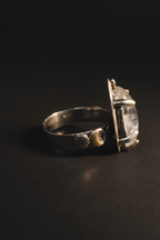 A silver-toned ring with a textured, hammered band and a large, irregular clear stone set in a rough, metallic frame, photographed on a dark, reflective surface.