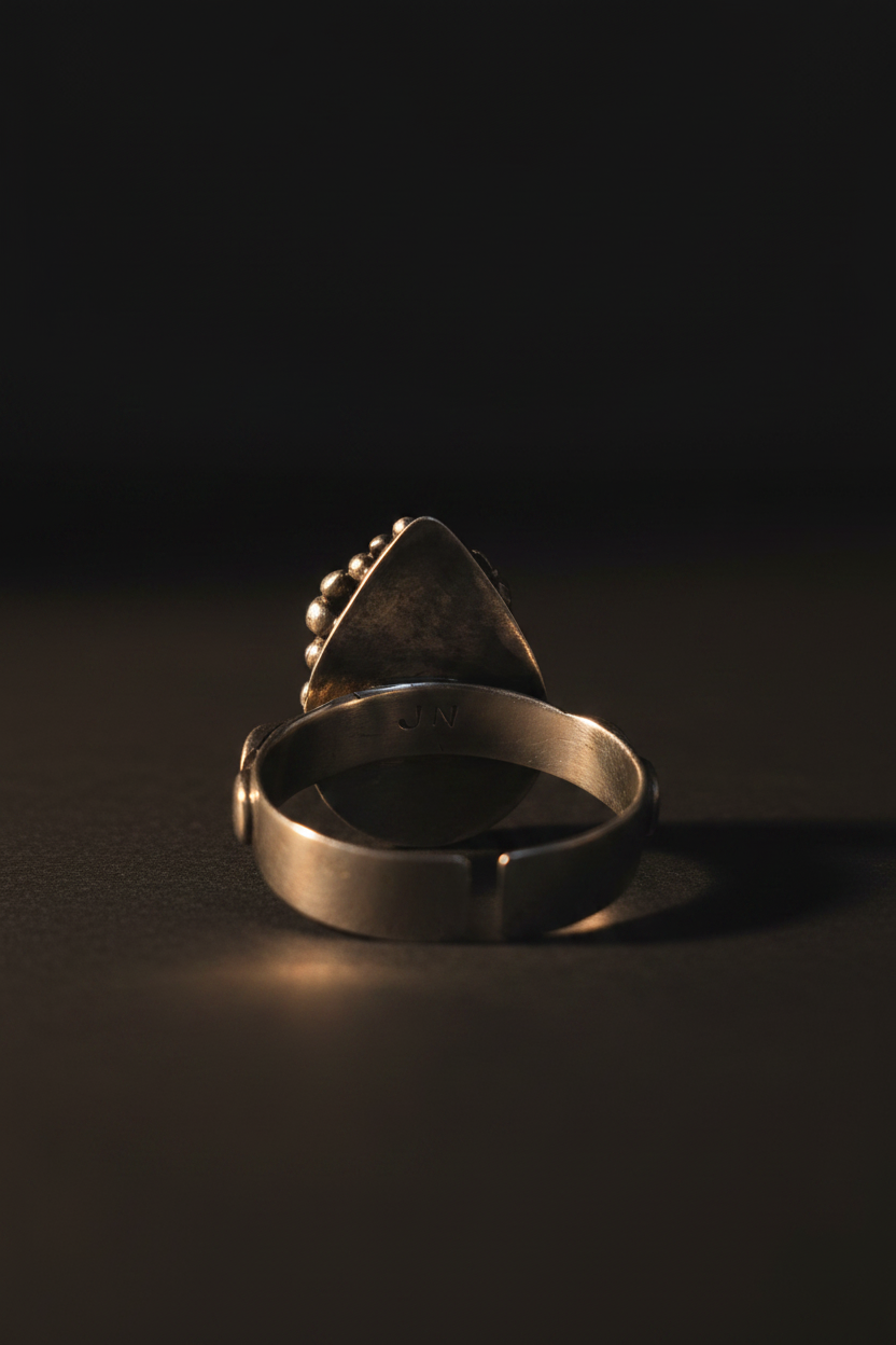 A vintage-style silver ring with a triangular top and small bead accents, photographed on a dark, reflective surface with dramatic lighting.