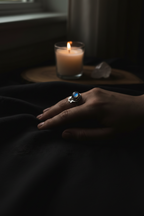 A silver ring with a round blue stone is worn on a person's hand resting on dark fabric, with a lit candle and crystal in the background.