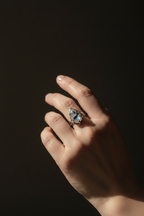 A close-up of a hand wearing a silver-toned ring with a marquise-cut iridescent blue stone, set against a dark, shadowed background.