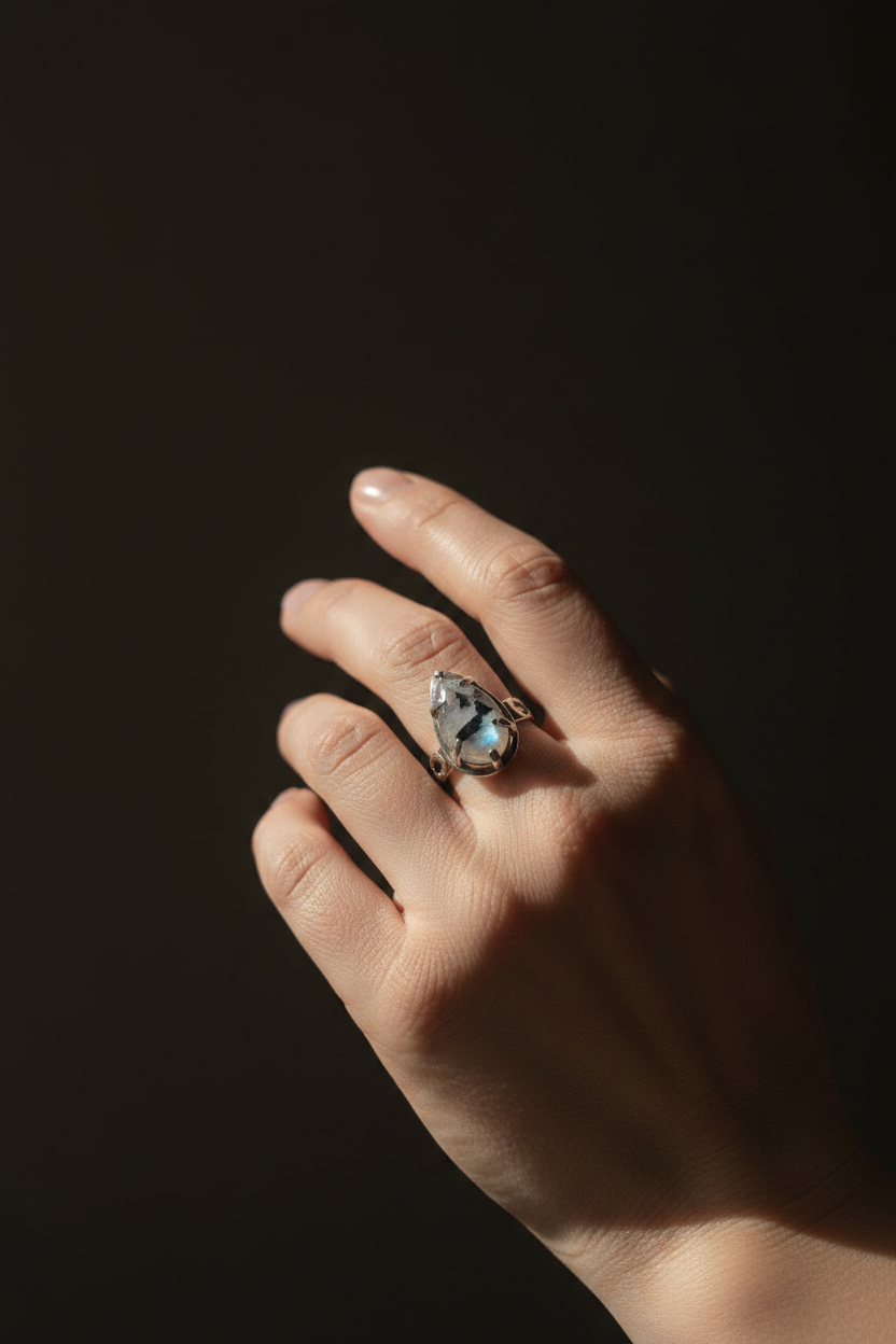 A close-up of a hand wearing a silver-toned ring with a marquise-cut iridescent blue stone, set against a dark, shadowed background.
