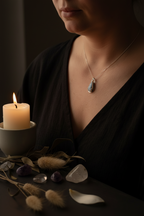 A close-up lifestyle shot of a woman wearing a silver pendant necklace with a blue-gray stone, styled with a lit candle, crystals, and dried grass on a dark table.