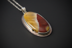 An oval pendant necklace featuring a split-color stone cabochon with yellow and dark red sections, set in a polished silver bezel on a matching silver chain, displayed against a dark gray background.