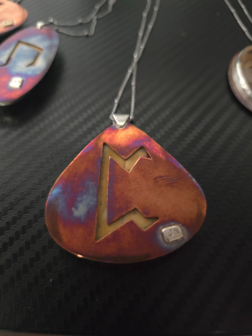 A close-up of a teardrop-shaped pendant with a guitar-pick silhouette, featuring a cutout design and a small square inset, shown on a dark textured background.