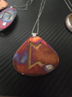 A close-up of a teardrop-shaped pendant with a guitar-pick silhouette, featuring a cutout design and a small square inset, shown on a dark textured background.