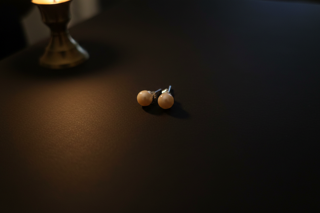 A pair of small stud earrings with round cream-colored beads on silver posts, resting on a dark surface under warm, dim lighting.