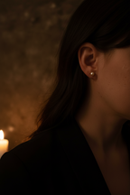A close-up side profile of a woman with dark hair wearing a small round gold stud earring, photographed in warm candlelit lighting against a dark textured background.