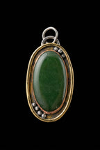 An oval pendant featuring a polished dark green cabochon stone set in a mixed metal frame with gold, copper, and silver tones, photographed on a dark textured background.