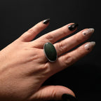 A close-up of a hand wearing a large oval ring with a dark green cabochon stone set in a gold-tone band.