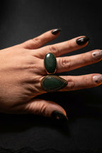 A close-up of a hand wearing two rings: a large oval dark green stone ring on the middle finger and a larger teardrop-shaped dark green stone ring on the ring finger, both with gold-toned settings.