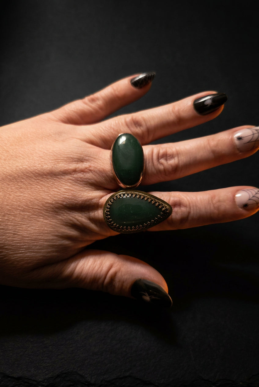 A close-up of a hand wearing two rings: a large oval dark green stone ring on the middle finger and a larger teardrop-shaped dark green stone ring on the ring finger, both with gold-toned settings.