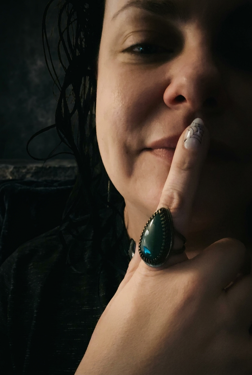 A close-up lifestyle shot of a woman wearing a large teardrop-shaped turquoise ring with a dark metal bezel and silver-toned French tip, resting her finger near her lips.