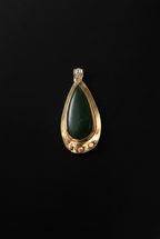 A teardrop-shaped pendant featuring a dark green stone in a gold-toned setting with a clear crystal bail, displayed on a dark background.
