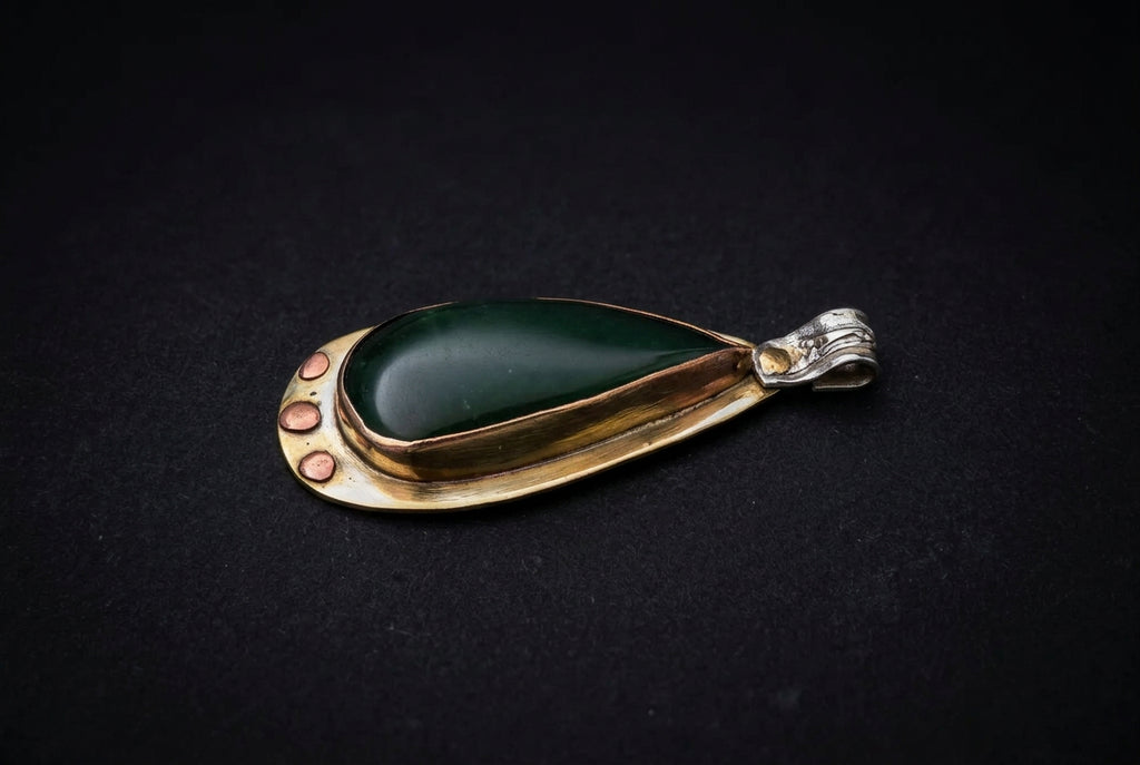 A teardrop-shaped pendant featuring a polished dark green cabochon stone set in a gold-toned metal frame with small gold dots on one side and a silver-toned bail on the other, displayed on a dark background.