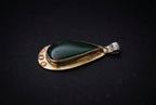 A teardrop-shaped pendant featuring a polished dark green cabochon stone set in a gold-toned metal frame with small gold dots on one side and a silver-toned bail on the other, displayed on a dark background.