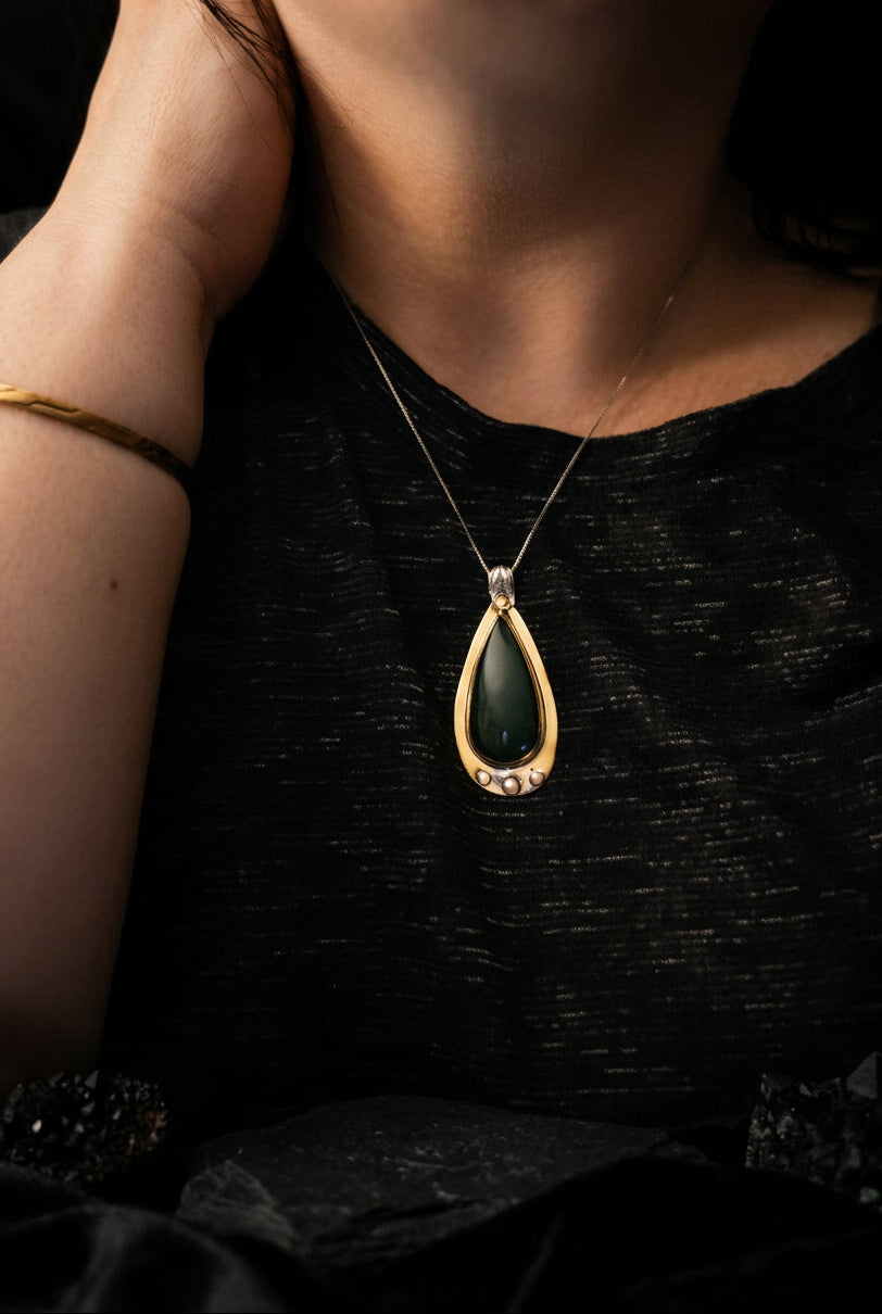 A close-up of a person wearing a silver-toned teardrop pendant necklace with a dark green stone, shown against a black textured top.