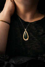A close-up of a person wearing a silver-toned teardrop pendant necklace with a dark green stone, shown against a black textured top.
