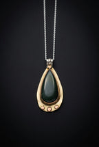 A teardrop-shaped pendant necklace with a dark green stone set in a gold-tone frame, suspended from a silver-tone chain against a black background.