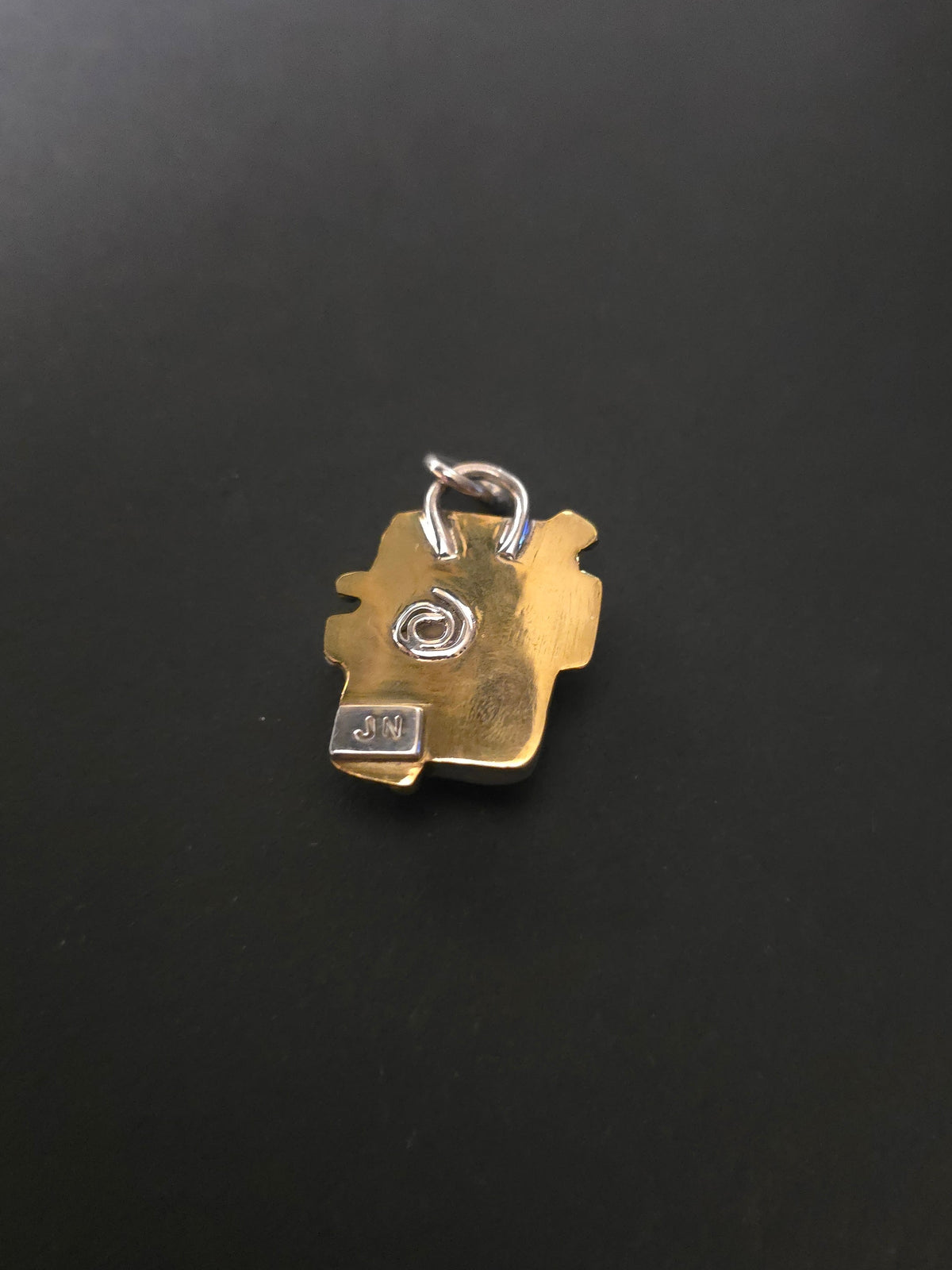 A small gold-toned pendant or charm with a silver ring attachment, photographed on a dark black surface.